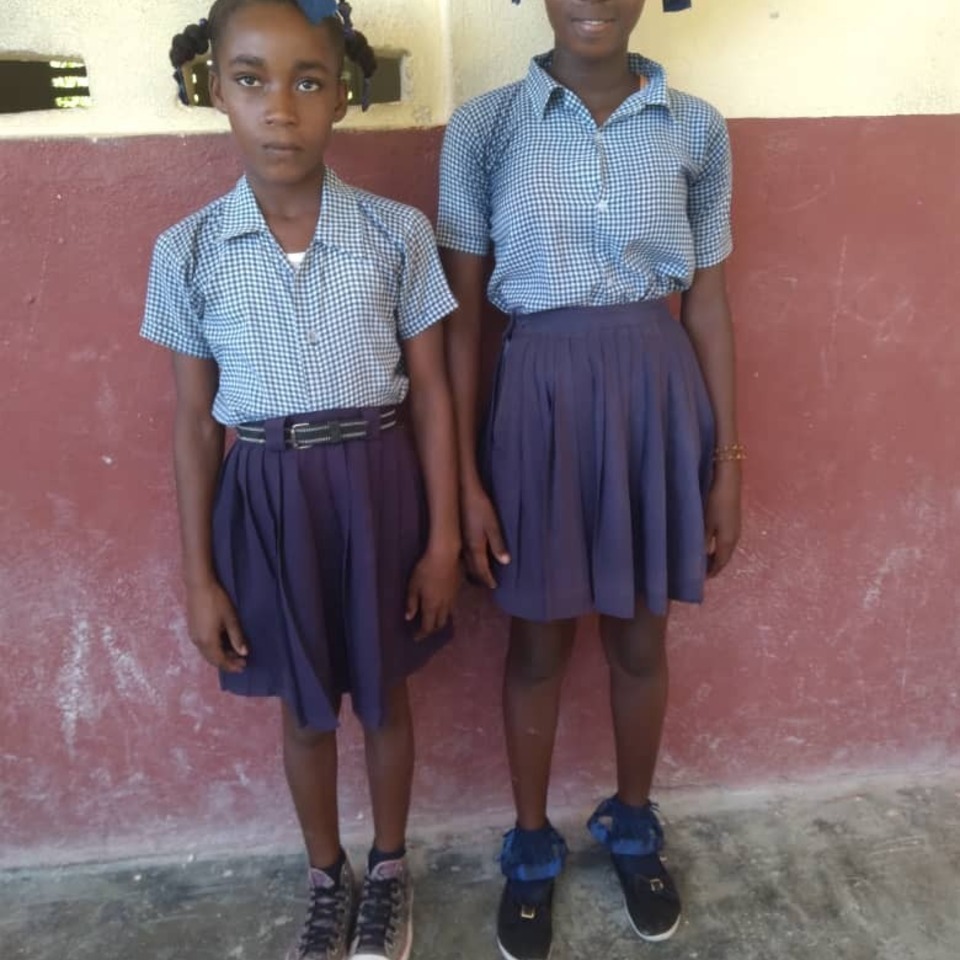 Two girls in blue shirts and skirts standing next to a wall.